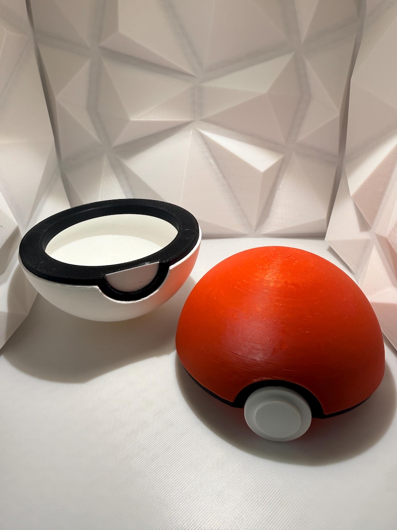May include: A red and white spherical container, resembling a cartoon Pokeball, is shown open and closed. The container is split in half, with a black rim and a white interior. The other half is a solid red sphere with a white button.