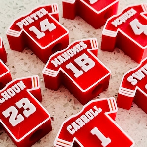 May include: Red jersey-shaped pencil sharpeners with white lettering and numbers. The names and numbers include Porter 14, Mahomes 15, McQueen 4, Swift 87, Carroll 1, and Calhoun 23. The sharpeners are arranged on a light-colored surface.