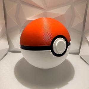 May include: A 3D-printed Pok&eacute; Ball, featuring a vibrant orange top and a white bottom, separated by a black band. A white circular button is centered on the front. The ball casts a shadow on the white surface.