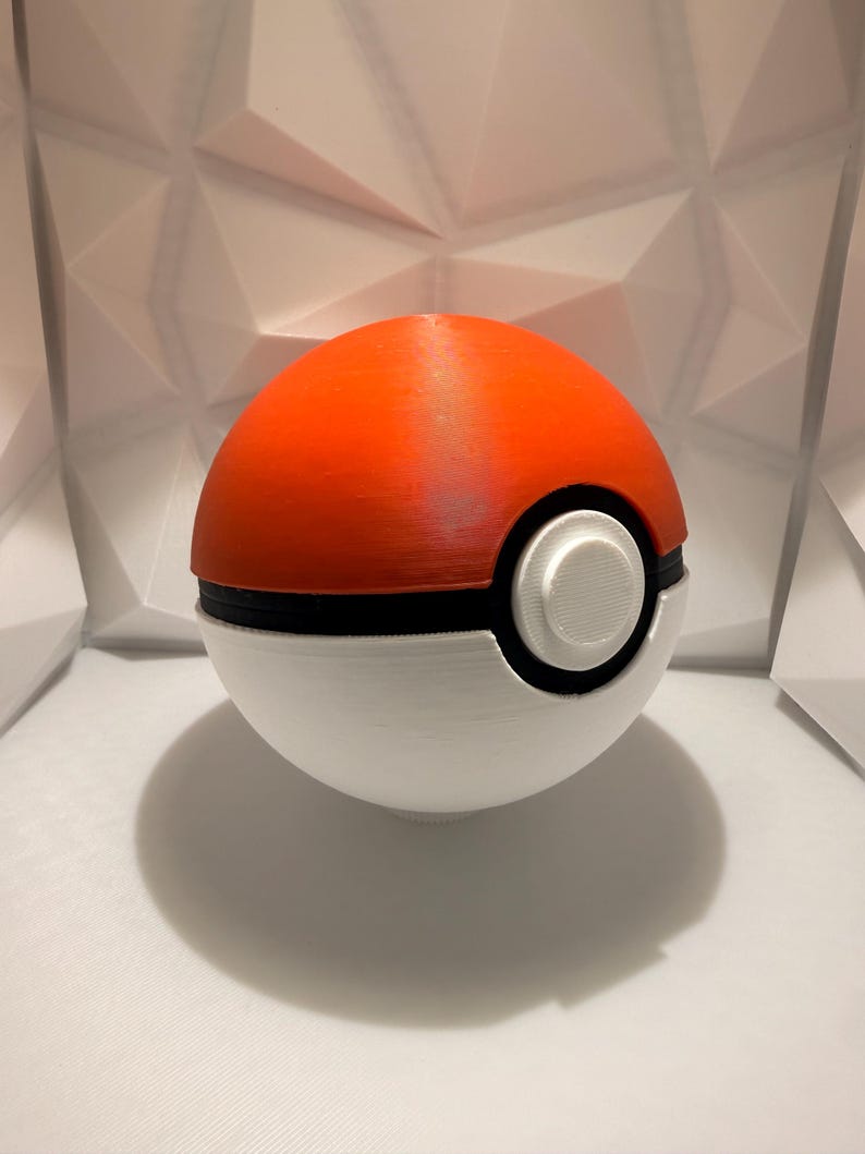 May include: A 3D-printed Pok&eacute; Ball, featuring a vibrant orange top and a white bottom, separated by a black band. A white circular button is centered on the front. The ball casts a shadow on the white surface.