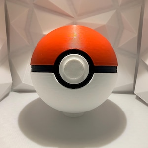 May include: A 3D-printed Poké Ball, a spherical object with a red upper half and a white lower half, separated by a black band. The center features a white circular button. The ball casts a shadow on the white surface.