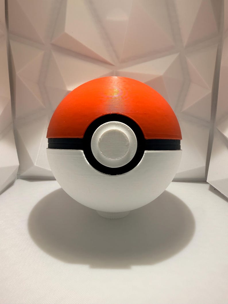 May include: A 3D-printed Pok&eacute; Ball, a spherical object with a red upper half and a white lower half, separated by a black band. The center features a white circular button. The ball casts a shadow on the white surface.
