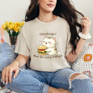 Könnte beinhalten: Eine Frau trägt ein beiges T-Shirt mit einer Comic-Katze, die einen Cheeseburger hält. Auf dem Shirt steht "Cheeseburger? Eher Cheese-PURRger!"