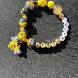 May include: A bracelet featuring yellow, gold, and black beads. The bracelet includes a charm of a yellow and black bee. The bracelet spells "BEE HAPPY" with gold and white letter beads. The bracelet is on a black surface.