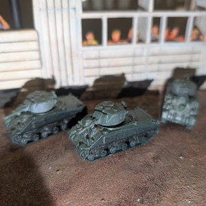 May include: Three olive green toy tanks with detailed treads and turrets are arranged on a brown surface. The tanks are miniature replicas, likely for play or display. The background includes a structure with figures.