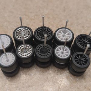May include: A collection of small, black model car tires with various wheel designs, including white and silver accents. Each tire has a silver pin inserted through the center. The tires are stacked in small groups.