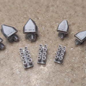 May include: A collection of small, gray, 3D-printed components. The assortment includes several cone-shaped pieces and rectangular pieces with multiple cylindrical holes. The items are arranged on a speckled, light brown surface.