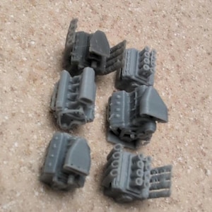 May include: A collection of six small, gray model engine components. The components have detailed features, including cylinders and other mechanical parts. The pieces are arranged on a light brown surface, suggesting a hobby or model-making theme.