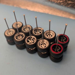 May include: A collection of miniature black tires with silver rims and long metal pins. Some tires have a red stripe around the edge. The tires are stacked in pairs, suggesting they are for a model car or similar craft project.