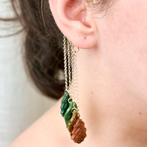 May include: Earrings with a gold chain and leaf design. The leaves are green, brown, and reddish-brown. The earrings are attached to the ear with a small gold hoop.