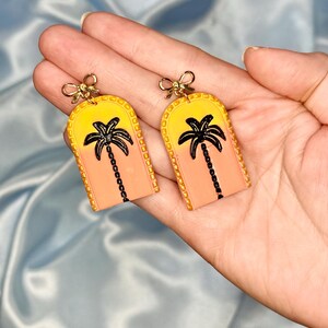 May include: A pair of arch-shaped dangle earrings featuring a yellow to peach gradient background with a black palm tree silhouette. Each earring is topped with a small gold-toned metal bow charm and has a textured border along the arched edge.