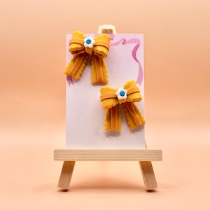 以下が含まれることがあります： 白と青の花の中心が付いた、黄色のリボン型のヘアクリップが2つ、白いカードに表示されています。カードは、桃色の背景に対して、小さな木製のイーゼルに立てかけられています。リボンは、テクスチャ加工されたレイヤーデザインになっています。