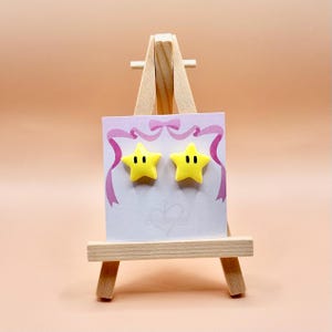 May include: A pair of yellow star-shaped stud earrings with black dot eyes, displayed on a white card with a pink ribbon design. The earrings are presented on a small wooden easel against a peach-colored background.