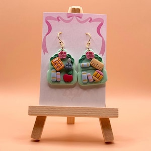 May include: A pair of dangle earrings featuring light green backpack-shaped charms. Each charm is decorated with miniature school supplies including a calculator, books, a red apple, scissors, a ruler, and a pencil, topped with a small pink flower.