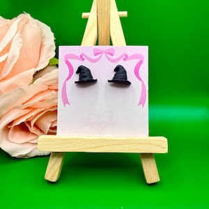 Puede incluir: Un par de pendientes de sombrero de bruja negros exhibidos en una tarjeta blanca con un diseño de lazo rosa. Los pendientes están sobre un pequeño caballete de madera sobre un fondo verde. Una gran rosa rosa es visible en el fondo.