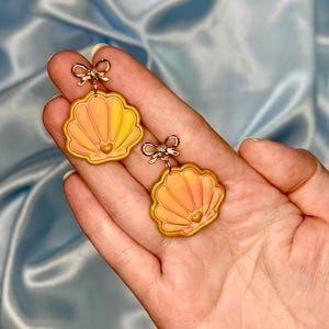 May include: A pair of dangle earrings featuring orange and yellow marbled seashell charms with gold-toned edges. Each shell has a small gold heart at the base and is suspended from a delicate gold-toned metal bow connector at the top.
