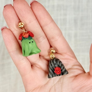 以下が含まれることがあります： ゴールドトーンの金具が付いた、お化けの形をしたチャームのぶら下がりイヤリング2点。1つのお化けは無地のライトグリーンで、頭部に赤とオレンジの花の房が付いています。もう1つのお化けは黒とグレーの縦縞模様で、赤い花が1つ付いています。
