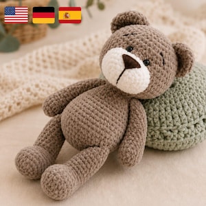 Pode incluir: Um urso de pelúcia de crochê feito à mão, em tons de castanho, com focinho branco e nariz castanho escuro. O urso está deitado sobre uma almofada verde de crochê. Os olhos do urso são pequenos e pretos. A estética geral é suave e aconchegante.