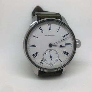 K&ouml;nnte beinhalten: Eine silberne Armbanduhr mit wei&szlig;em Zifferblatt, schwarzen r&ouml;mischen Ziffern und einer kleinen Sekundenscheibe. Die Uhr hat ein dunkelgr&uuml;nes Lederarmband und eine strukturierte Krone. Auf dem Zifferblatt steht "SR. UETZSCHNER".