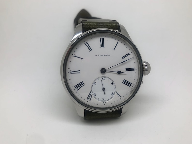 K&ouml;nnte beinhalten: Eine silberne Armbanduhr mit wei&szlig;em Zifferblatt, schwarzen r&ouml;mischen Ziffern und einer kleinen Sekundenscheibe. Die Uhr hat ein dunkelgr&uuml;nes Lederarmband und eine strukturierte Krone. Auf dem Zifferblatt steht "SR. UETZSCHNER".
