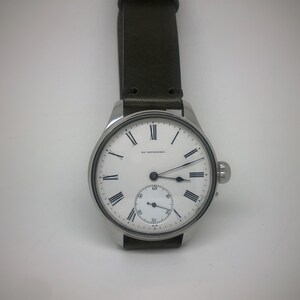 K&ouml;nnte beinhalten: Eine silberfarbene Armbanduhr mit wei&szlig;em Zifferblatt und schwarzen r&ouml;mischen Ziffern. Die Uhr hat ein kleines Sekunden-Zifferblatt und ein dunkelgr&uuml;nes Lederarmband. Auf dem Zifferblatt steht "R.F. WETZORKES".