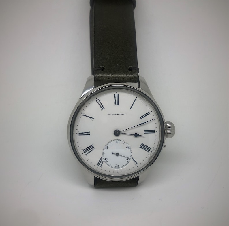 K&ouml;nnte beinhalten: Eine silberfarbene Armbanduhr mit wei&szlig;em Zifferblatt und schwarzen r&ouml;mischen Ziffern. Die Uhr hat ein kleines Sekunden-Zifferblatt und ein dunkelgr&uuml;nes Lederarmband. Auf dem Zifferblatt steht "R.F. WETZORKES".