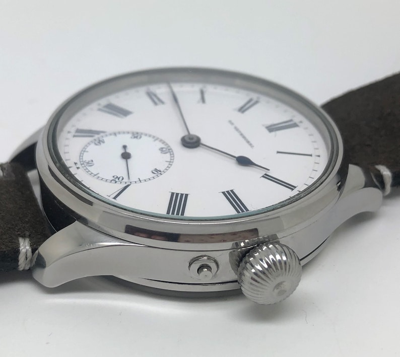 K&ouml;nnte beinhalten: Nahaufnahme einer silbernen Armbanduhr mit wei&szlig;em Zifferblatt, schwarzen r&ouml;mischen Ziffern und einer kleinen Sekundenanzeige. Die Uhr hat ein braunes Wildlederarmband und eine strukturierte Krone.