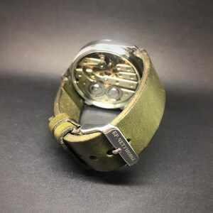 K&ouml;nnte beinhalten: Eine Armbanduhr mit silberfarbenem Geh&auml;use und sichtbarem mechanischem Uhrwerk. Sie hat ein gr&uuml;nes Lederarmband mit einer silbernen Schnalle. Auf der Schnalle steht "БР. ЧЕТУНОВЫ".