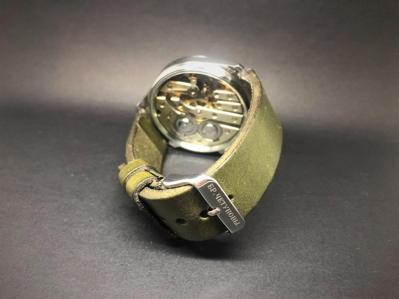 K&ouml;nnte beinhalten: Eine Armbanduhr mit silberfarbenem Geh&auml;use und sichtbarem mechanischem Uhrwerk. Sie hat ein gr&uuml;nes Lederarmband mit einer silbernen Schnalle. Auf der Schnalle steht "БР. ЧЕТУНОВЫ".