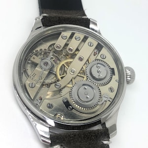 K&ouml;nnte beinhalten: Eine silberfarbene Armbanduhr mit transparentem Zifferblatt, das die komplizierten internen Zahnr&auml;der und Mechanismen zeigt. Die Uhr hat ein schwarzes Lederarmband und eine Aufzugskrone. Das Zifferblatt zeigt r&ouml;mische Ziffern.