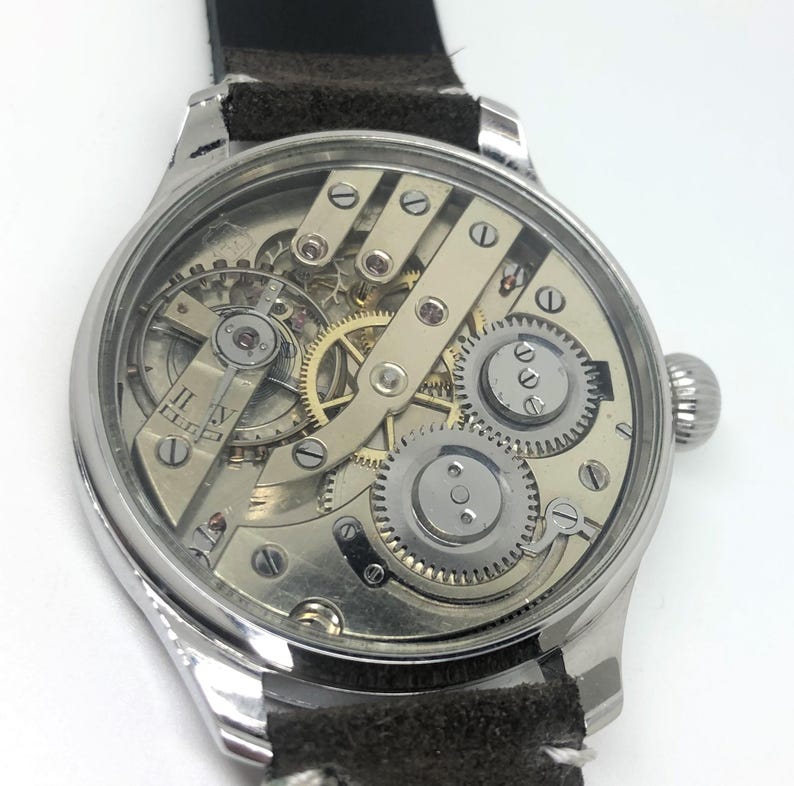 K&ouml;nnte beinhalten: Eine silberfarbene Armbanduhr mit transparentem Zifferblatt, das die komplizierten internen Zahnr&auml;der und Mechanismen zeigt. Die Uhr hat ein schwarzes Lederarmband und eine Aufzugskrone. Das Zifferblatt zeigt r&ouml;mische Ziffern.
