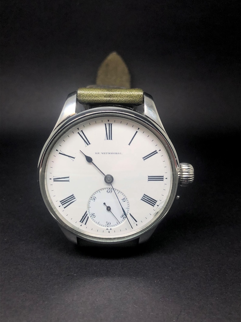 K&ouml;nnte beinhalten: Eine silberfarbene Armbanduhr mit wei&szlig;em Zifferblatt und schwarzen r&ouml;mischen Ziffern. Die Uhr hat ein kleines Sekunden-Zifferblatt und ein gr&uuml;nes Lederarmband. Auf dem Zifferblatt steht "SF. NETHERHOL".