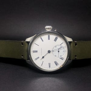 K&ouml;nnte beinhalten: Eine silberfarbene Armbanduhr mit wei&szlig;em Zifferblatt, schwarzen r&ouml;mischen Ziffern und einem kleinen Hilfszifferblatt. Die Uhr hat ein gr&uuml;nes Lederarmband. Die Uhr ist vor einem dunklen Hintergrund platziert.