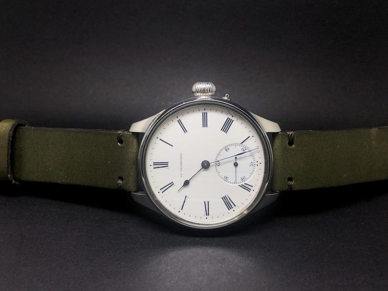 K&ouml;nnte beinhalten: Eine silberfarbene Armbanduhr mit wei&szlig;em Zifferblatt, schwarzen r&ouml;mischen Ziffern und einem kleinen Hilfszifferblatt. Die Uhr hat ein gr&uuml;nes Lederarmband. Die Uhr ist vor einem dunklen Hintergrund platziert.