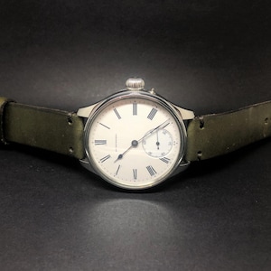 K&ouml;nnte beinhalten: Eine silberfarbene Armbanduhr mit wei&szlig;em Zifferblatt und schwarzen r&ouml;mischen Ziffern. Die Uhr hat ein kleines Sekunden-Zifferblatt und eine silberne Krone. Sie hat ein gr&uuml;nes Lederarmband mit einer silbernen Schnalle.
