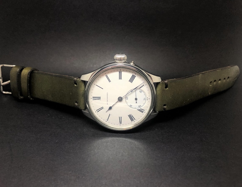 K&ouml;nnte beinhalten: Eine silberfarbene Armbanduhr mit wei&szlig;em Zifferblatt und schwarzen r&ouml;mischen Ziffern. Die Uhr hat ein kleines Sekunden-Zifferblatt und eine silberne Krone. Sie hat ein gr&uuml;nes Lederarmband mit einer silbernen Schnalle.