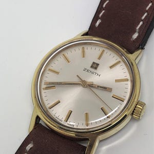 Può includere: Un orologio Zenith dorato con quadrante color crema e lancette e indici dorati. L'orologio ha un cinturino in pelle marrone con cuciture bianche. Il quadrante mostra il logo Zenith e le parole "T Swiss Made T".