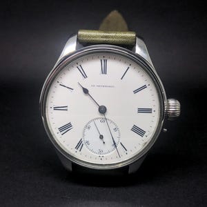 K&ouml;nnte beinhalten: Eine silberne Vintage-Armbanduhr mit wei&szlig;em Zifferblatt, schwarzen r&ouml;mischen Ziffern und einem kleinen Sekundenzeiger. Die Uhr hat ein gr&uuml;nes Lederarmband und eine strukturierte Aufzugskrone. Auf dem Zifferblatt steht "HE NETHERWOOD".