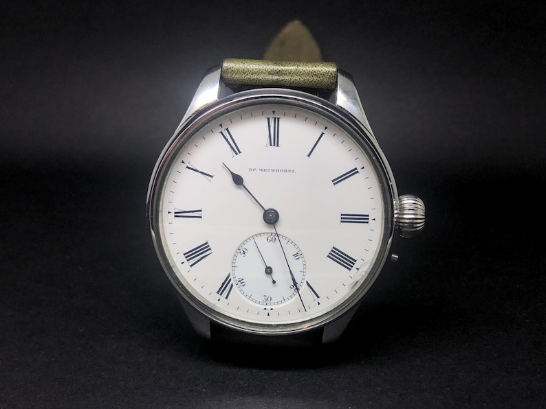 K&ouml;nnte beinhalten: Eine silberne Vintage-Armbanduhr mit wei&szlig;em Zifferblatt, schwarzen r&ouml;mischen Ziffern und einem kleinen Sekundenzeiger. Die Uhr hat ein gr&uuml;nes Lederarmband und eine strukturierte Aufzugskrone. Auf dem Zifferblatt steht "HE NETHERWOOD".