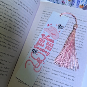 May include: Pink and white bookmark with a pink tassel. The bookmark features a pink cowboy hat with a black and white cow print band, a pink horseshoe, and the text "Wear This Hat, Cowboy".