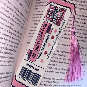 May include: Pink bookmark with the text "THE SMUTTY BOOK CLUB" and "ADMITS ONE." The bookmark has a pink tassel and is set against the backdrop of an open book. The design includes hearts and a stamp-like graphic.