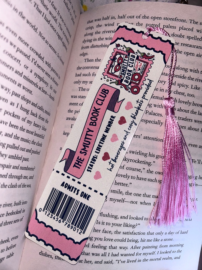 May include: Pink bookmark with the text "THE SMUTTY BOOK CLUB" and "ADMITS ONE." The bookmark has a pink tassel and is set against the backdrop of an open book. The design includes hearts and a stamp-like graphic.