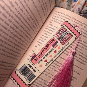 May include: A pink and cream bookmark with the text "THE SMUTTY BOOK CLUB" and "ADMITS ONE." It features a pink tassel and a barcode. The bookmark is placed on an open book.