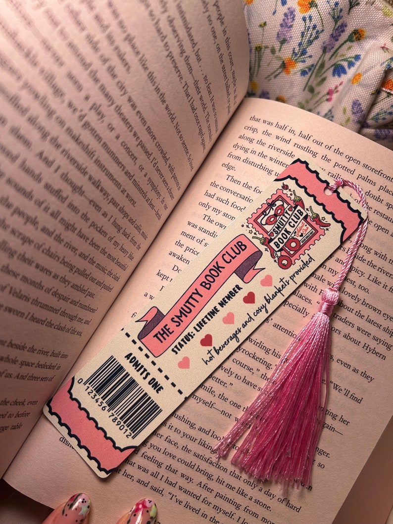 May include: A pink and cream bookmark with the text "THE SMUTTY BOOK CLUB" and "ADMITS ONE." It features a pink tassel and a barcode. The bookmark is placed on an open book.