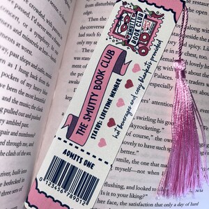 May include: A pink and white bookmark with the text "THE SMUTTY BOOK CLUB" and "ADMITS ONE." It features a pink tassel and a decorative border. The bookmark is in a book.