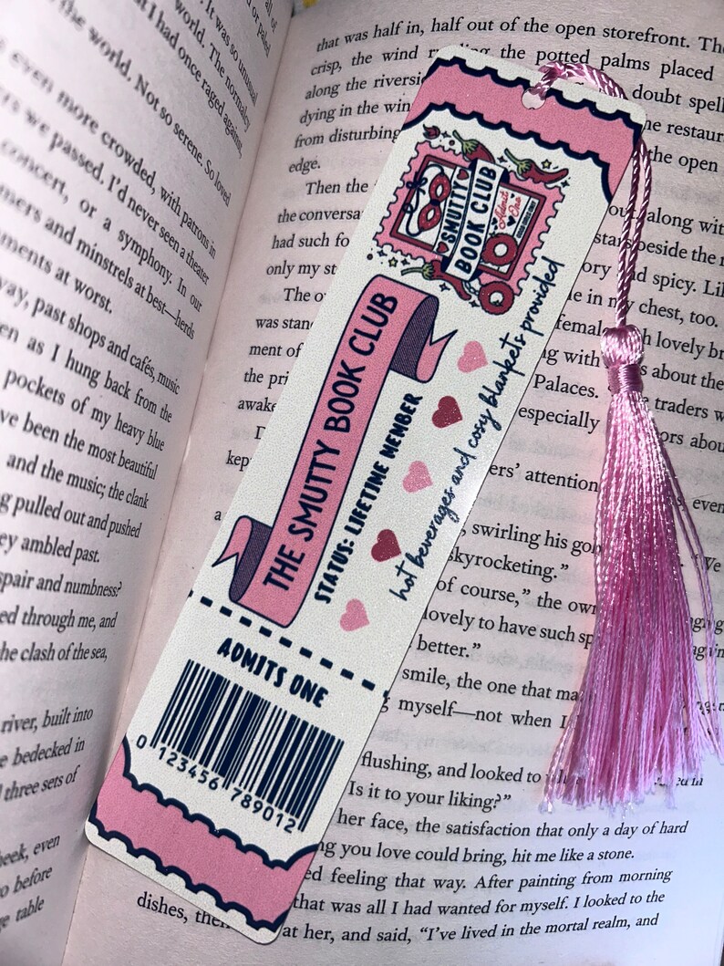 May include: A pink and white bookmark with the text "THE SMUTTY BOOK CLUB" and "ADMITS ONE." It features a pink tassel and a decorative border. The bookmark is in a book.