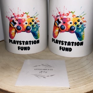 May include: Two white ceramic money jars with a colorful paint splatter design of a video game controller. The text "Playstation Fund" is printed on each jar.