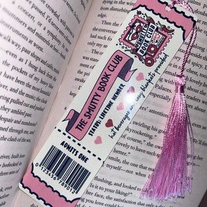 May include: A pink and white bookmark with the text "THE SMUTTY BOOK CLUB" and "ADMITS ONE." It features a pink tassel and is placed in an open book. The bookmark has a barcode and a stamp design.