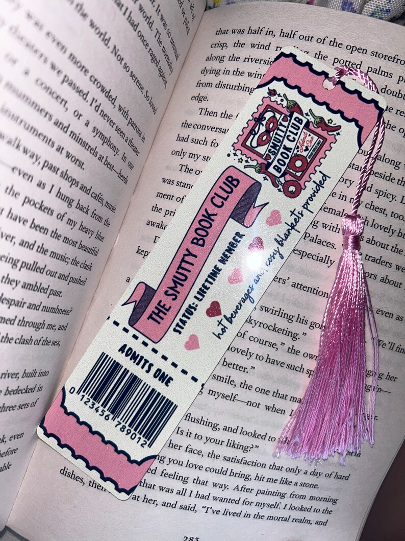 May include: A pink and white bookmark with the text "THE SMUTTY BOOK CLUB" and "ADMITS ONE." It features a pink tassel and is placed in an open book. The bookmark has a barcode and a stamp design.