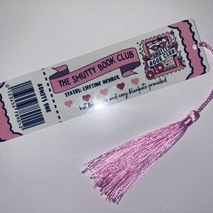May include: A bookmark with a pink tassel featuring the text "THE SMUTTY BOOK CLUB". The bookmark has a ticket design with a barcode and the words "ADMITS ONE". The text "STATUS: LIFETIME MEMBER" is also visible.